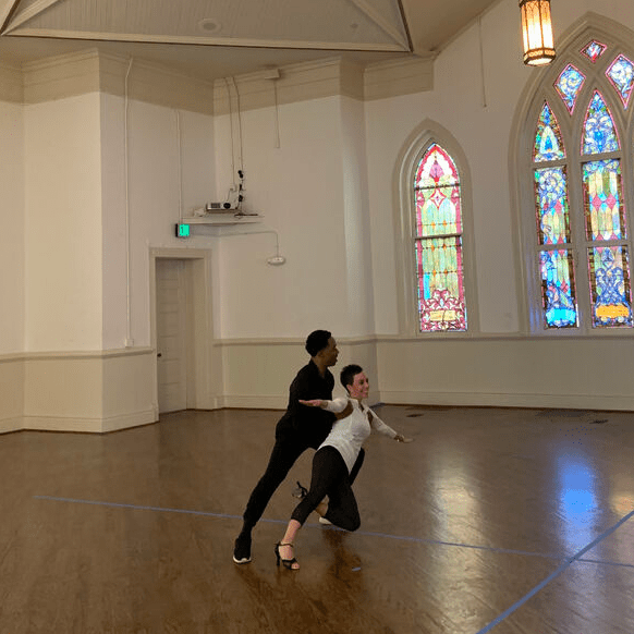 Kim Henderson and Maurice Mouzon, Jr. practice ballroom dance in a spacious building with a stained-glass window